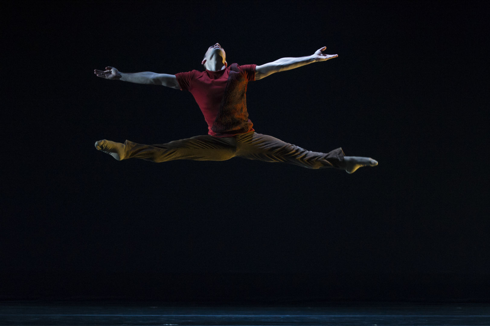 A man in a red shirt and ballet tights in the middle of a ballet jump.